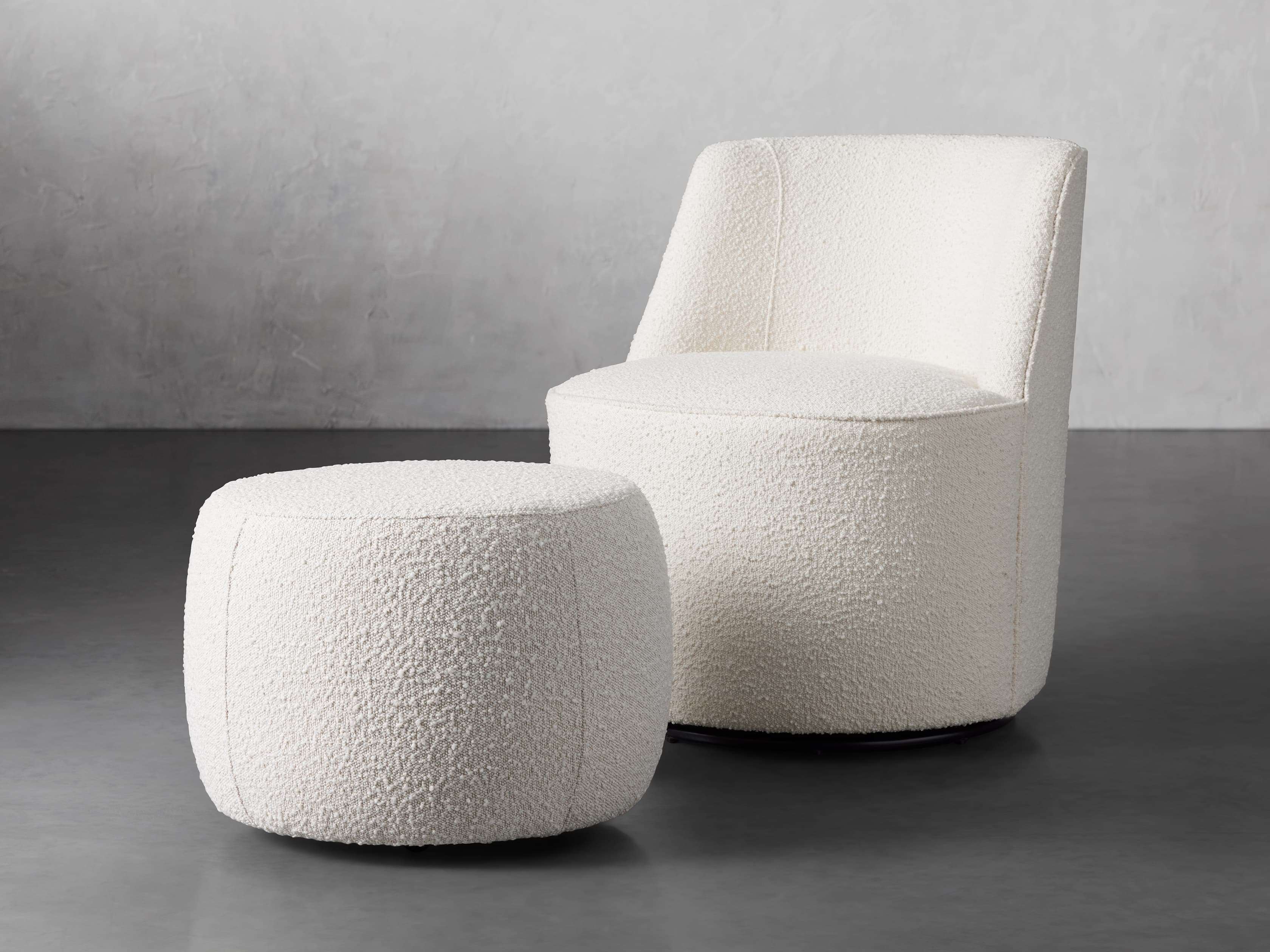 A modern upholstered ottoman in a natural color with a rounded soft design, displayed on a smooth gray floor in a minimalist setting.
