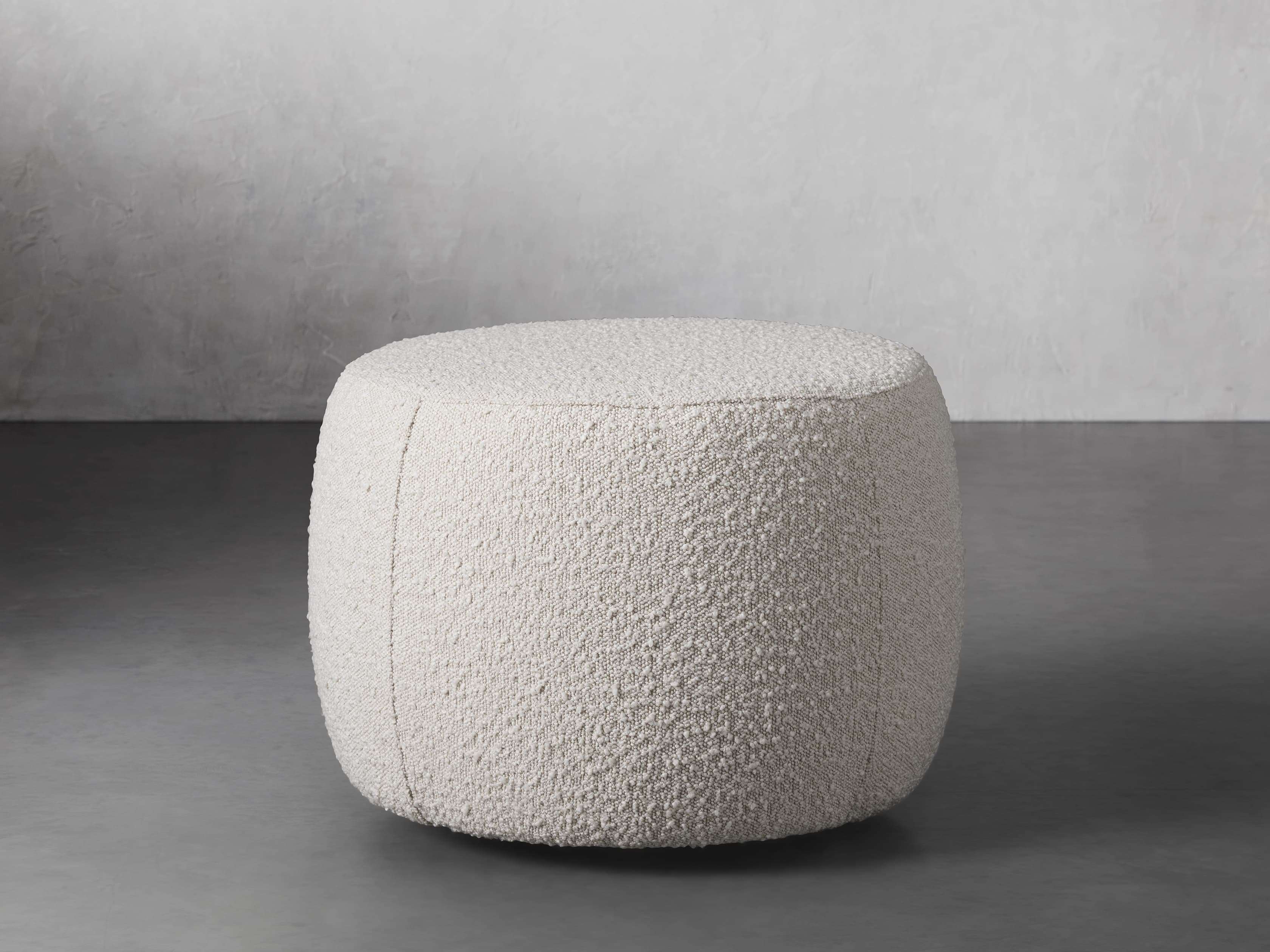 A rounded ottoman with a textured, natural off-white fabric, set on a smooth gray floor against a light wall.