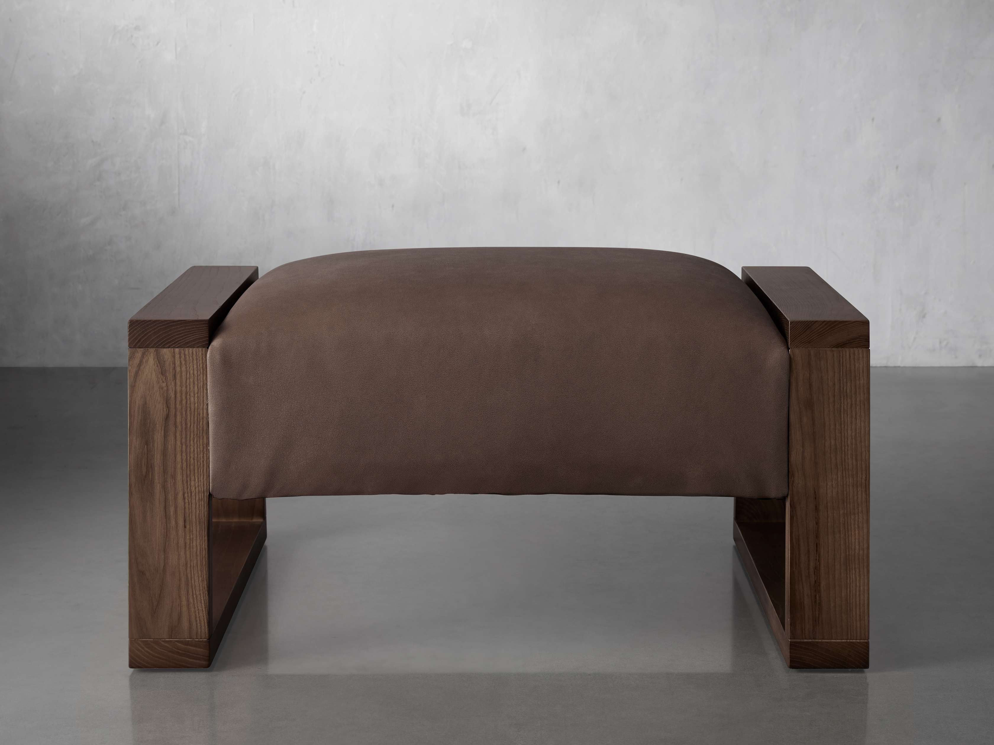 Brown leather ottoman with a rectangular shape, supported by medium wood frame legs, set against a neutral backdrop.