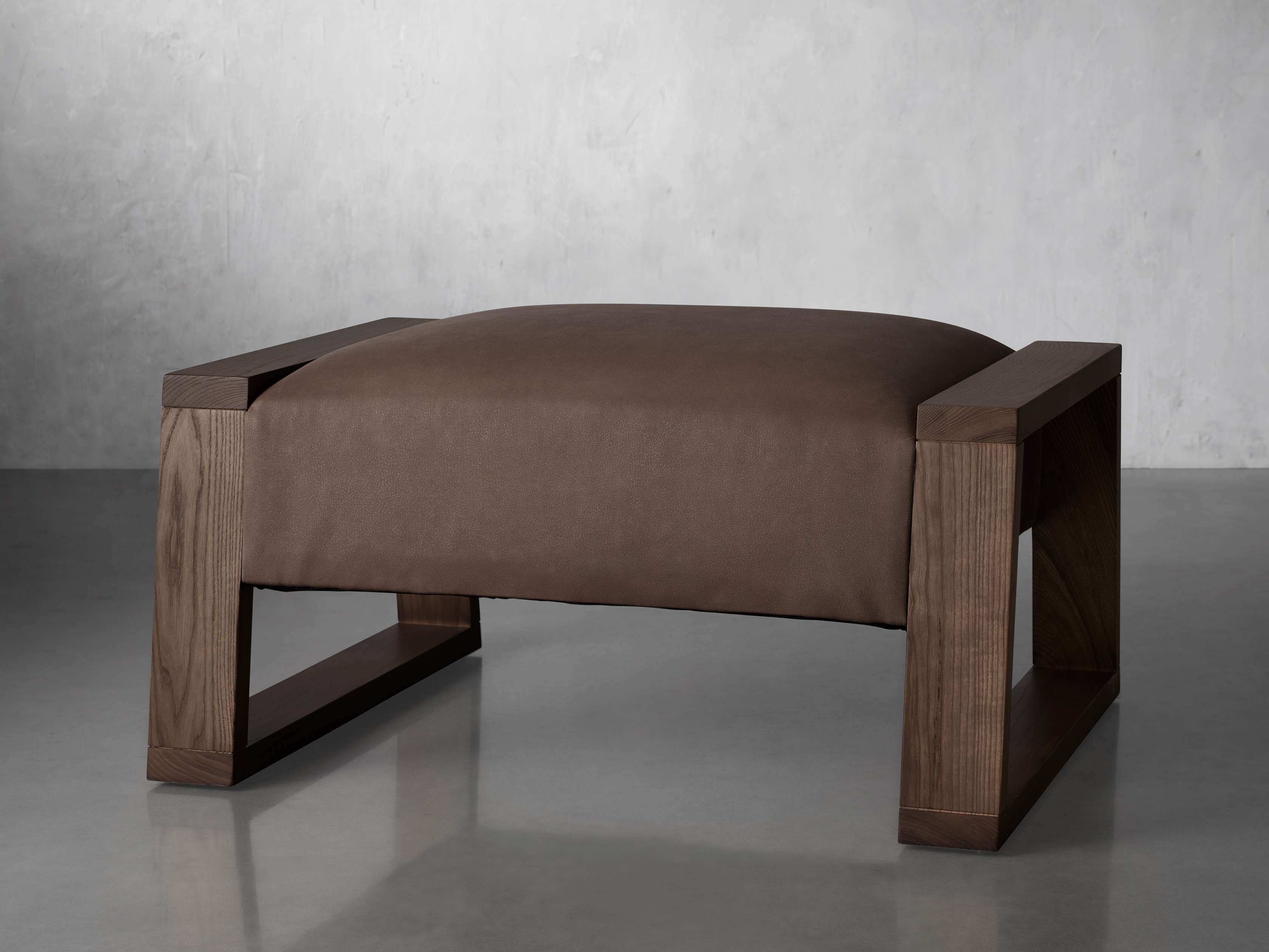 A brown leather ottoman with a rectangular wooden frame featuring visible wood grain and an open design, placed on a gray surface.