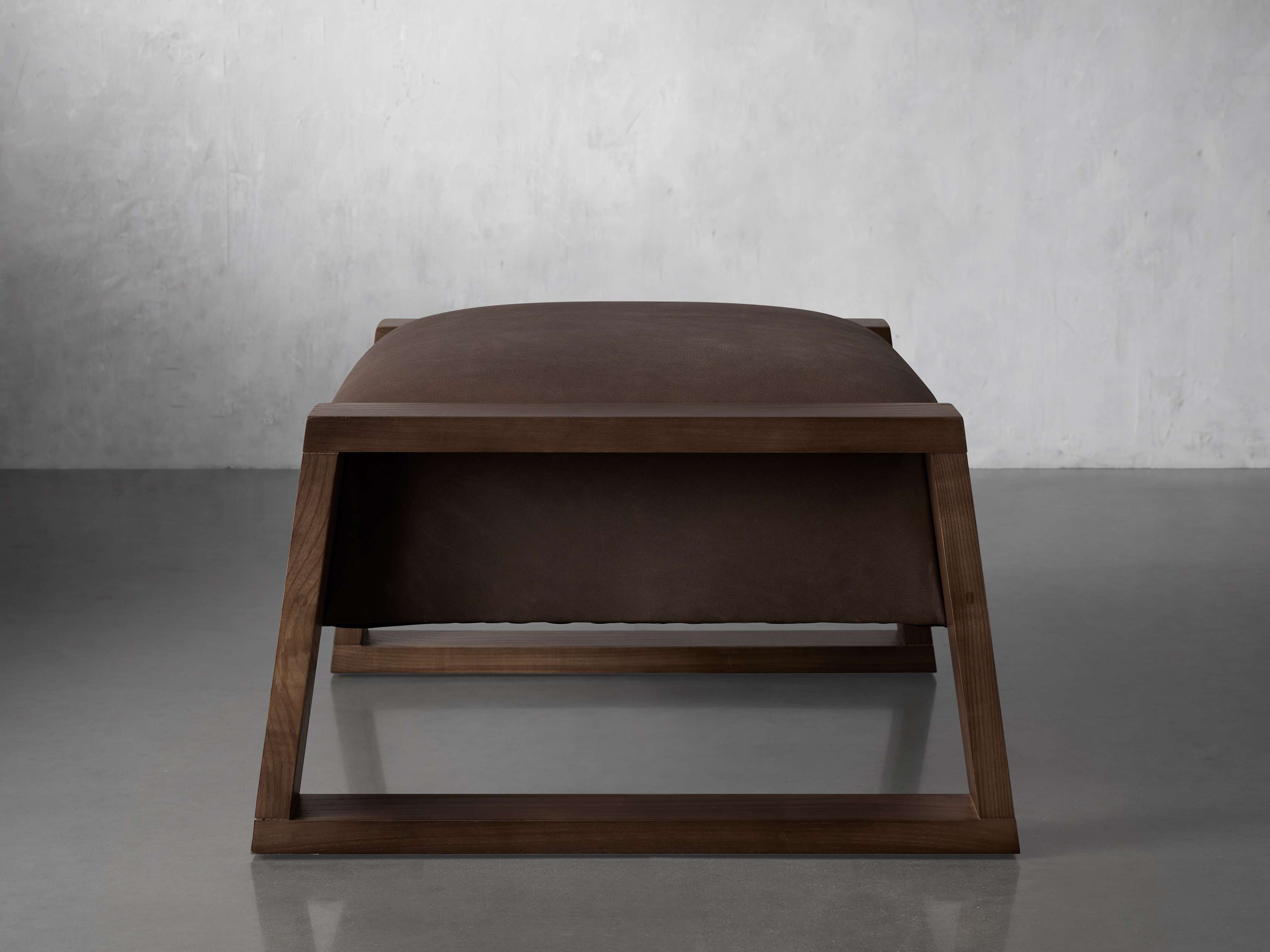 A brown leather upholstered ottoman with an angled wooden frame, displayed in a minimalist interior setting.