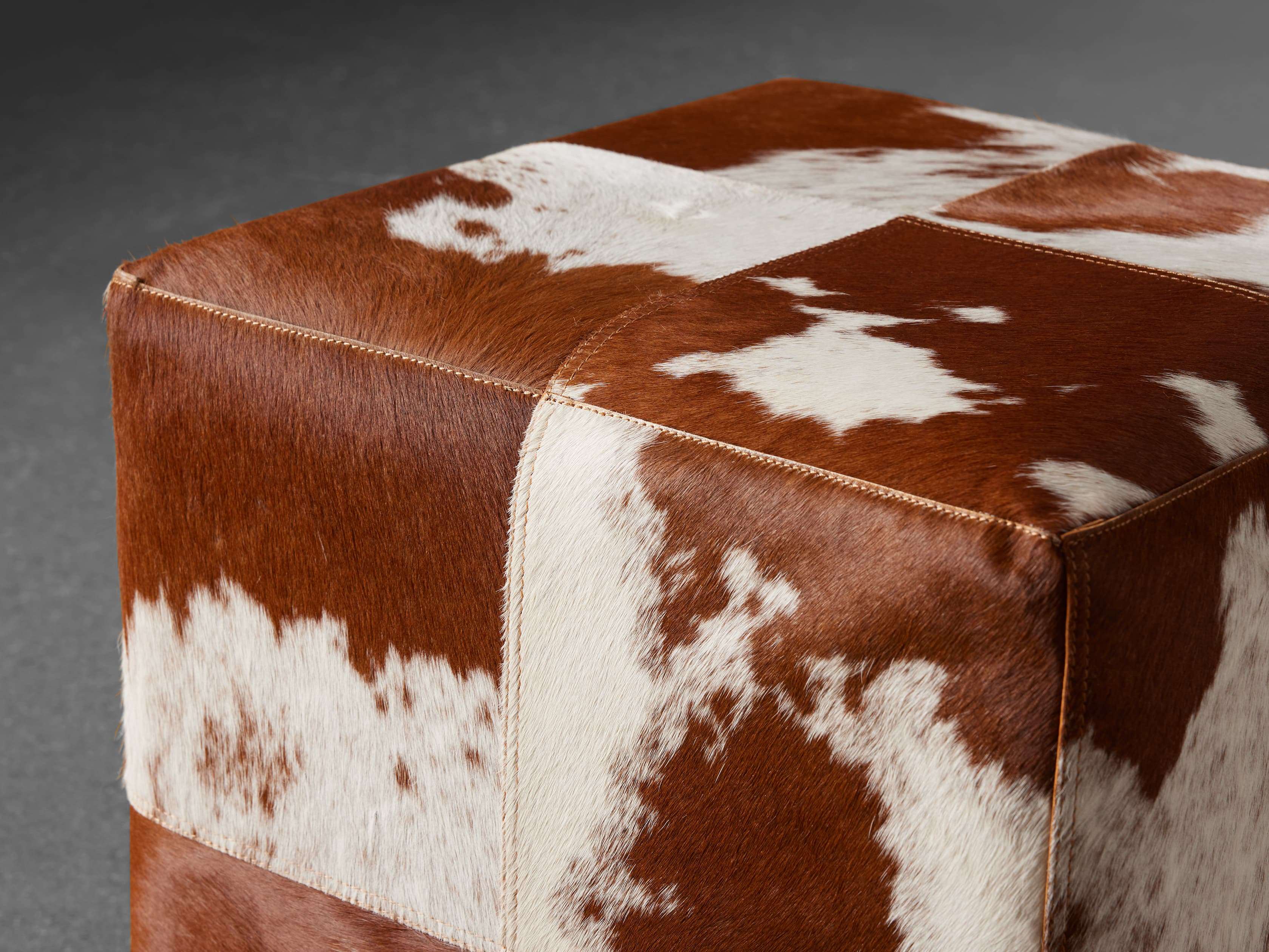 A square cowhide pouf with a patchwork pattern in shades of brown and white, showcasing natural textures and a rustic charm.