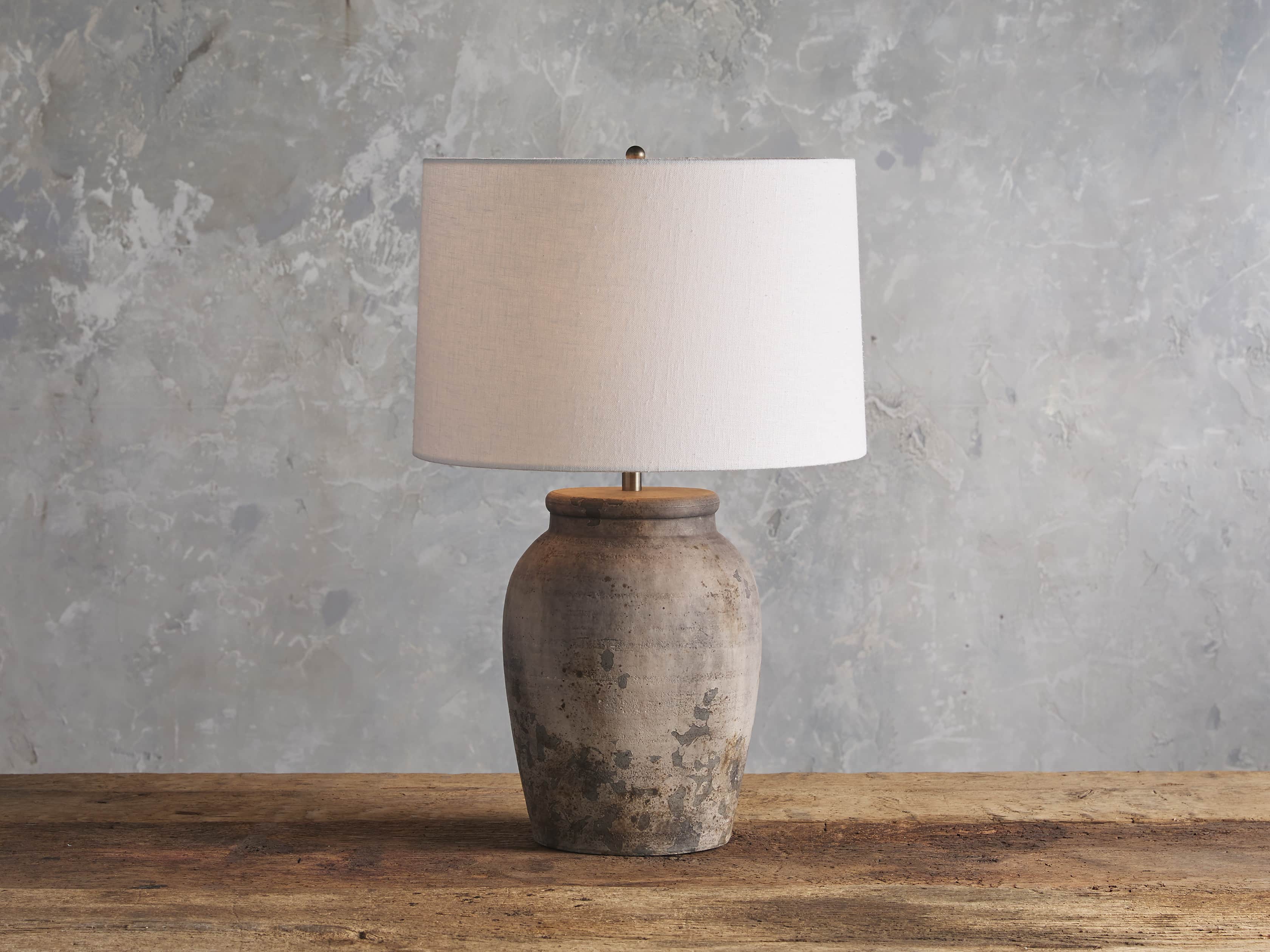 A ceramic table lamp with an antiqued gray finish, featuring an urn-shaped base and a white drum shade, placed on a wooden surface.