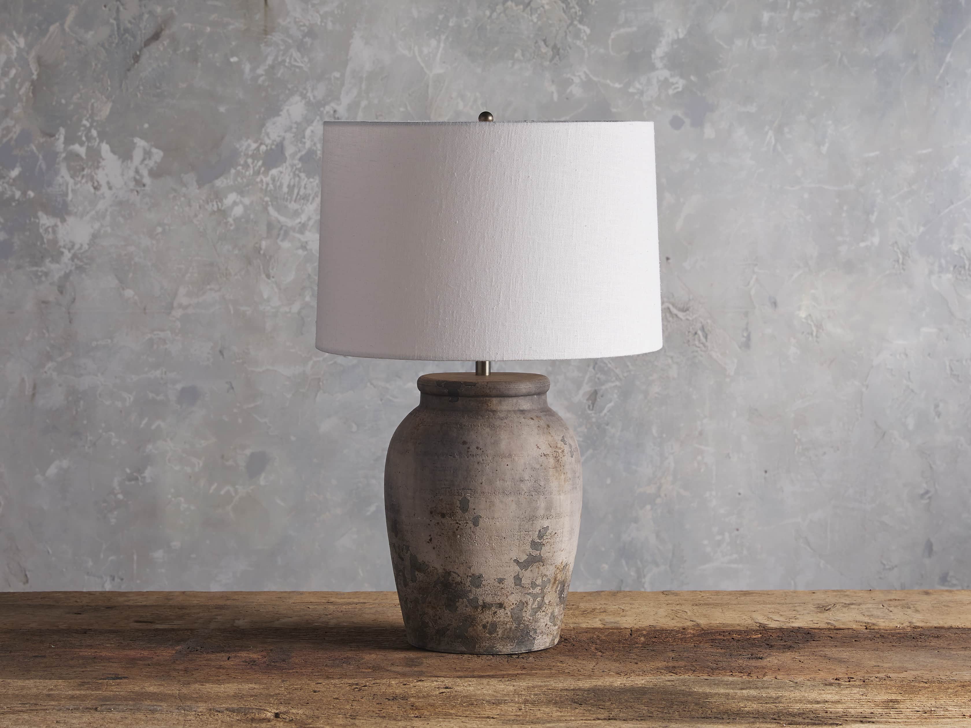 A ceramic table lamp with an antiqued gray finish, featuring an urn-shaped base and a white drum shade, placed on a wooden surface.