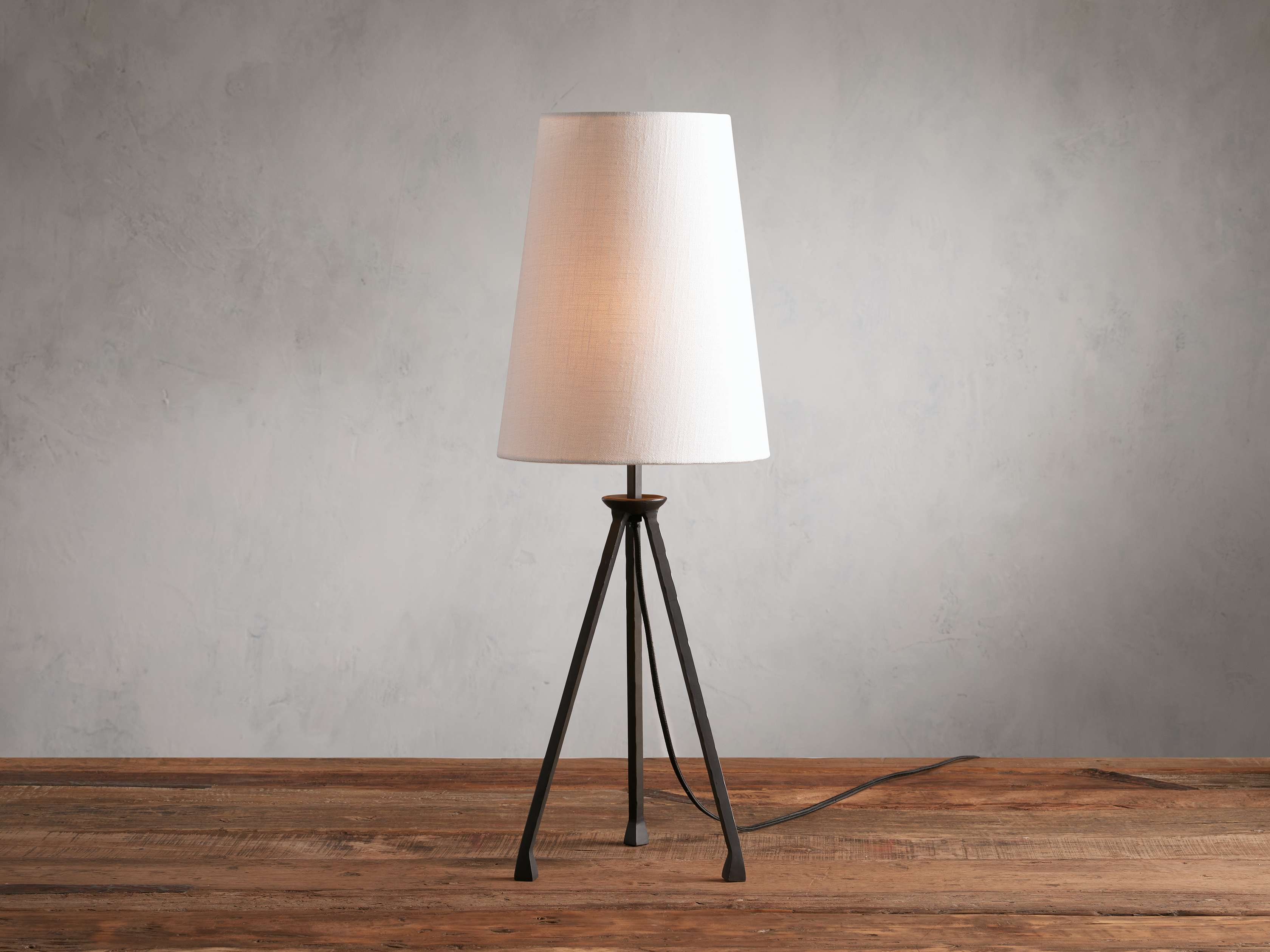 A black iron table lamp with a tripod base and a tall, elongated white linen shade, placed on a wooden surface against a neutral background.