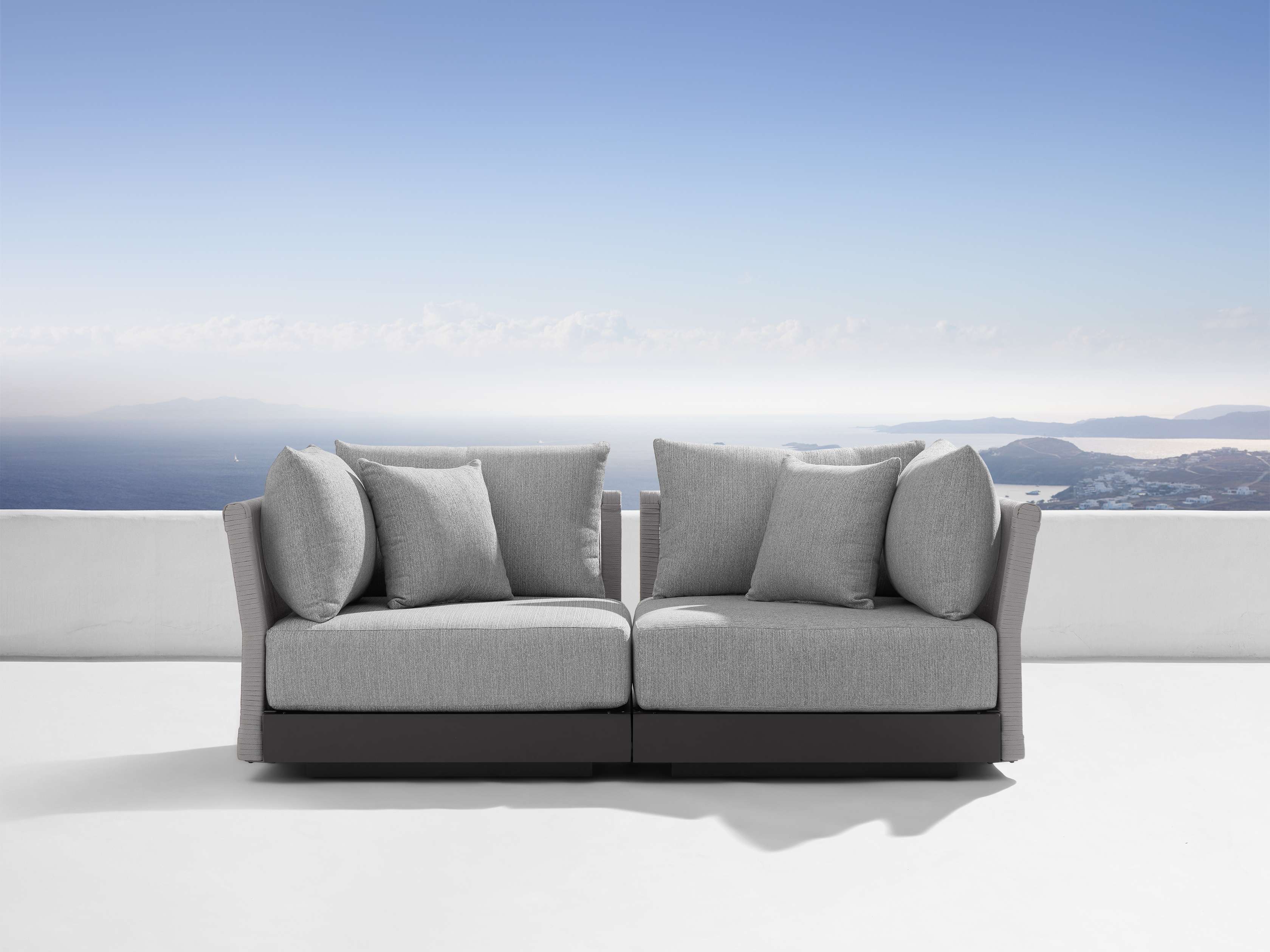 A two-piece modular outdoor sofa with gray cushions and a minimalist design, set against a clear blue sky and ocean background.