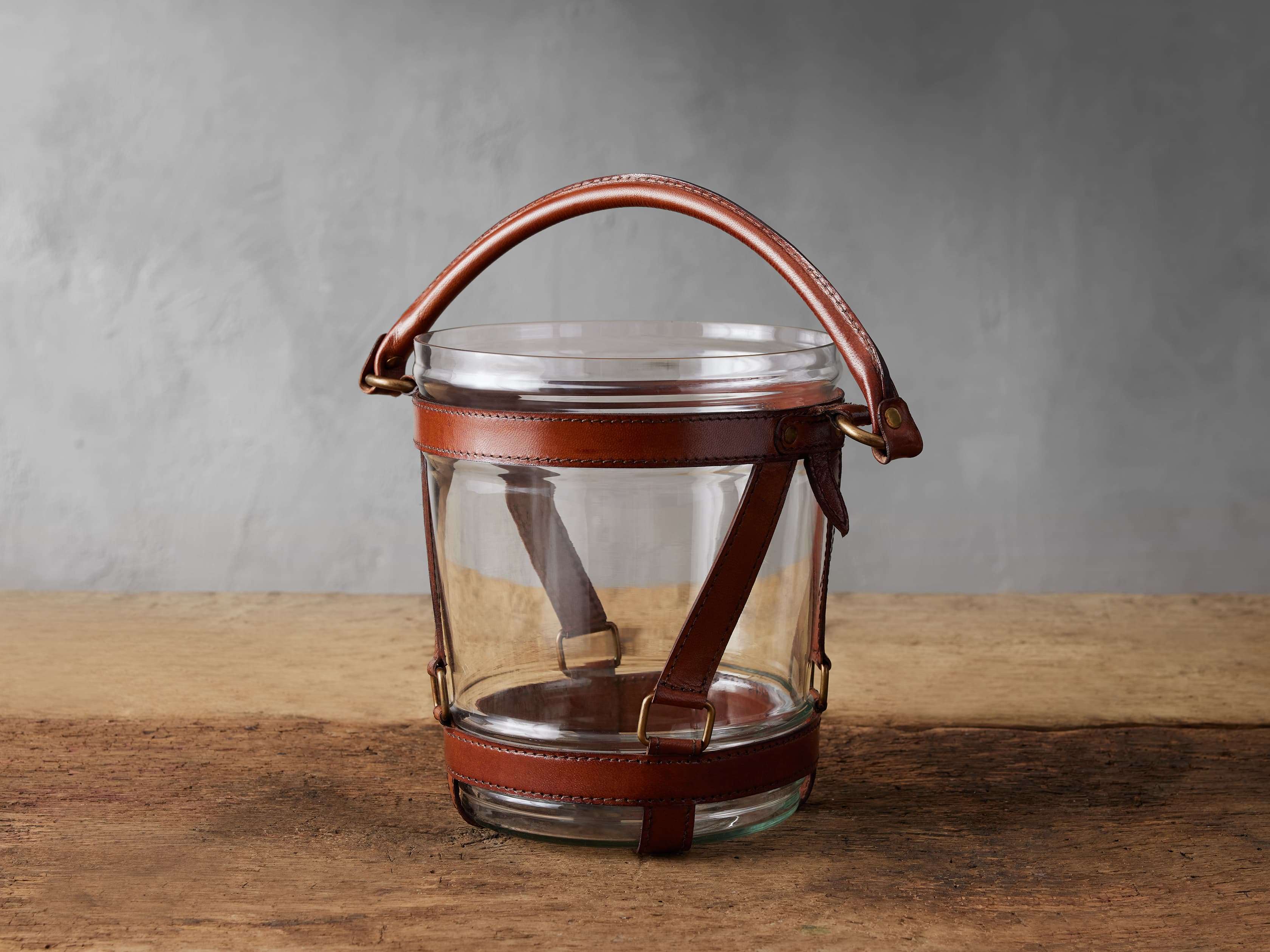 A round glass ice bucket with brown leather straps and a handle, featuring brass accents, sitting on a wooden surface.