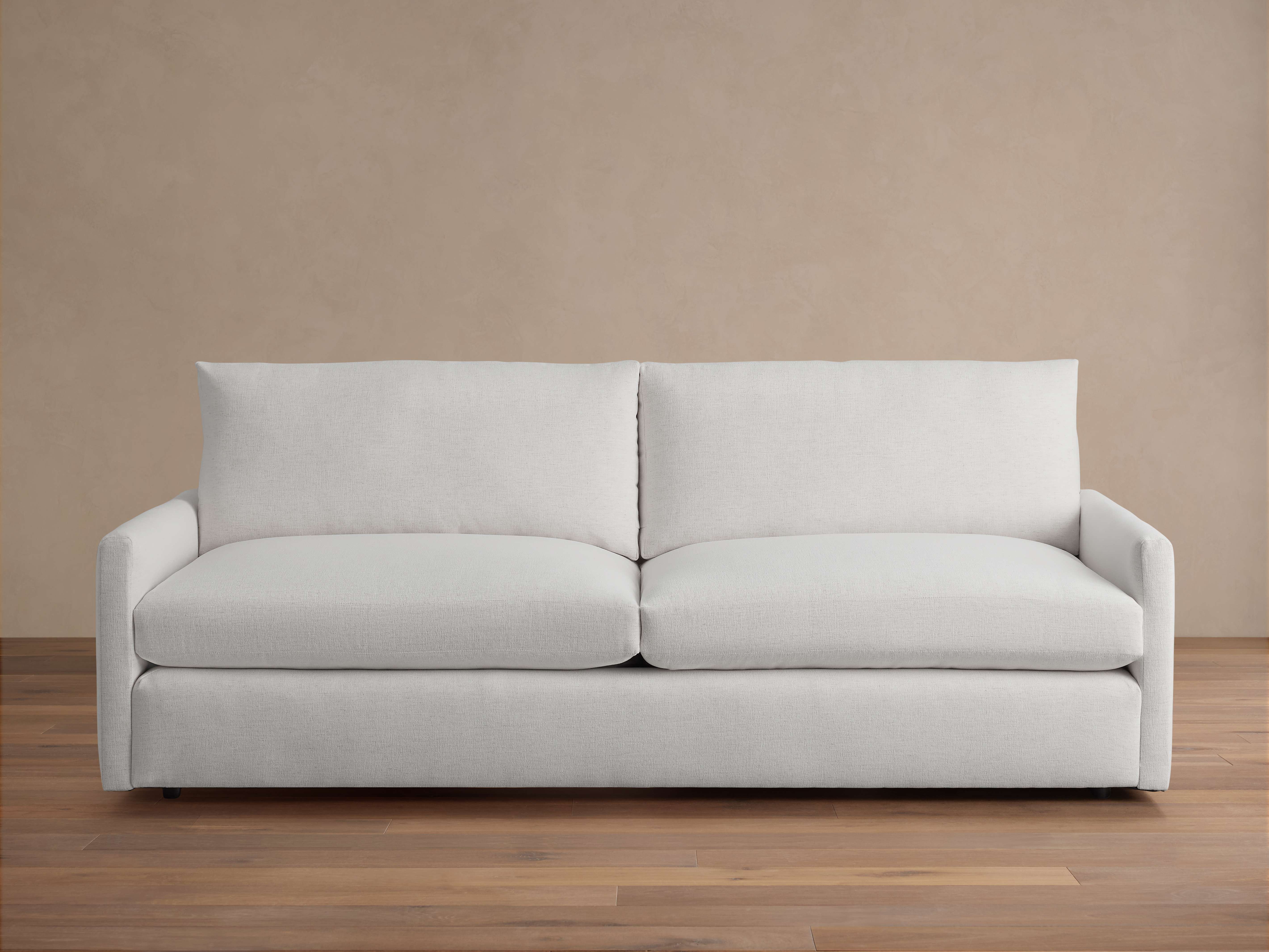 A light-colored Kipton Sofa with plush seat and back cushions; features a simple, modern design on a wooden floor.