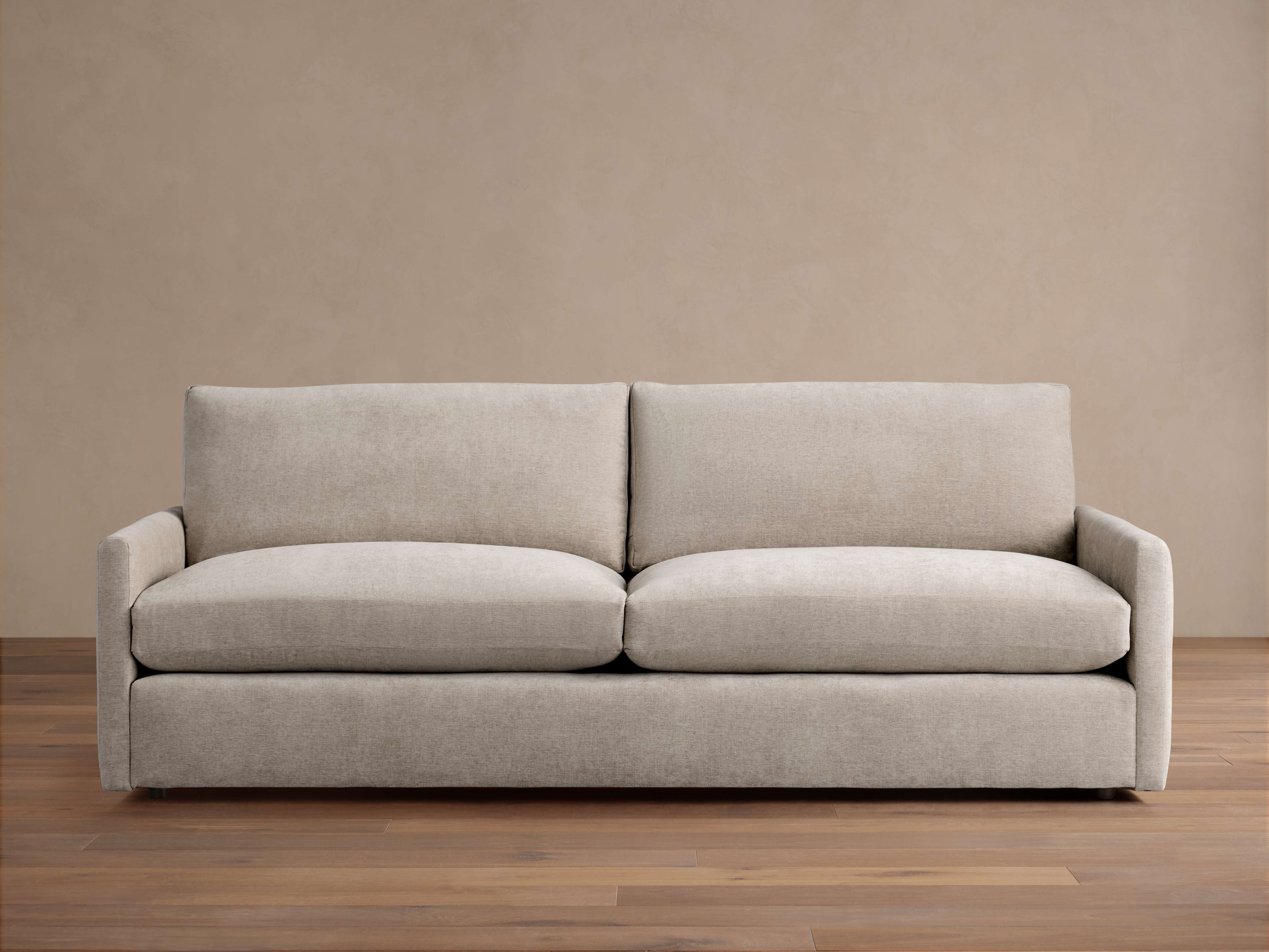 Kipton Slim Arm Sofa, upholstered in light beige fabric, with two seat and back cushions, slim arms, against a tan wall on a wooden floor.