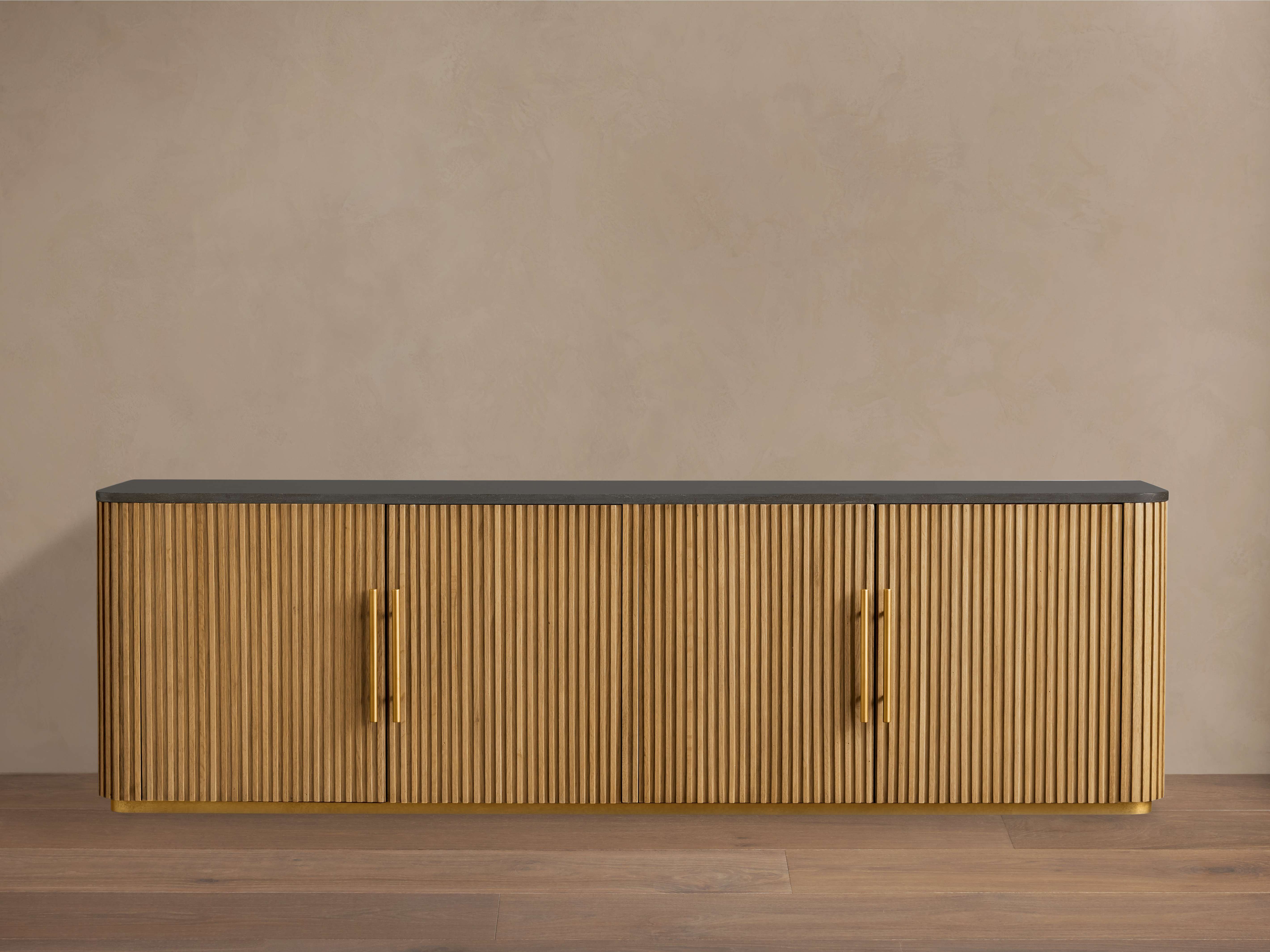 Finnley Media Console in Honey finish with fluted wood design, black top, and brass-finished pulls, sitting on wooden floor.