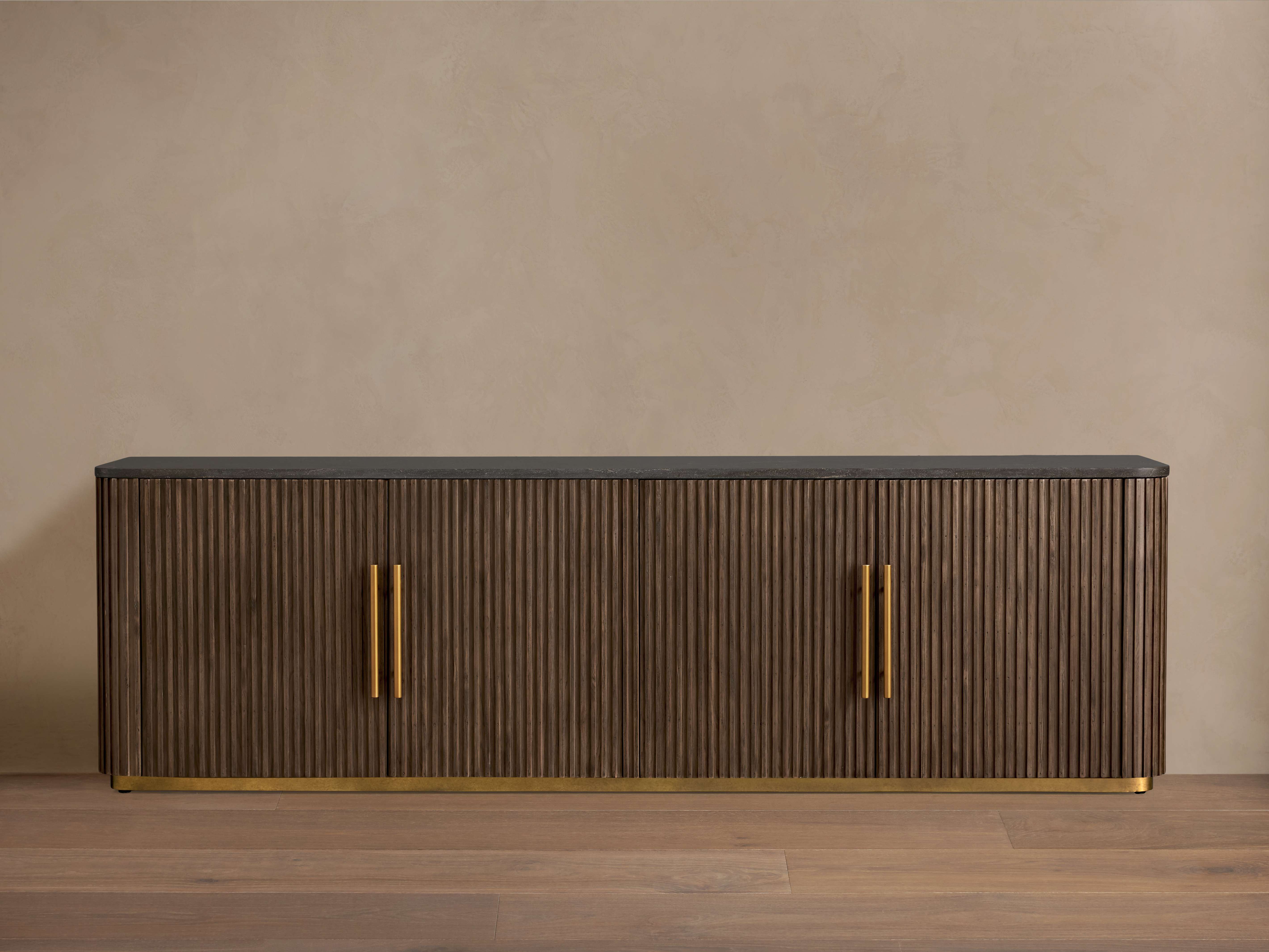 Finnley Media Console with vertical ribbed wood front, sleek black top, and brass handles. It rests on a light wooden floor.