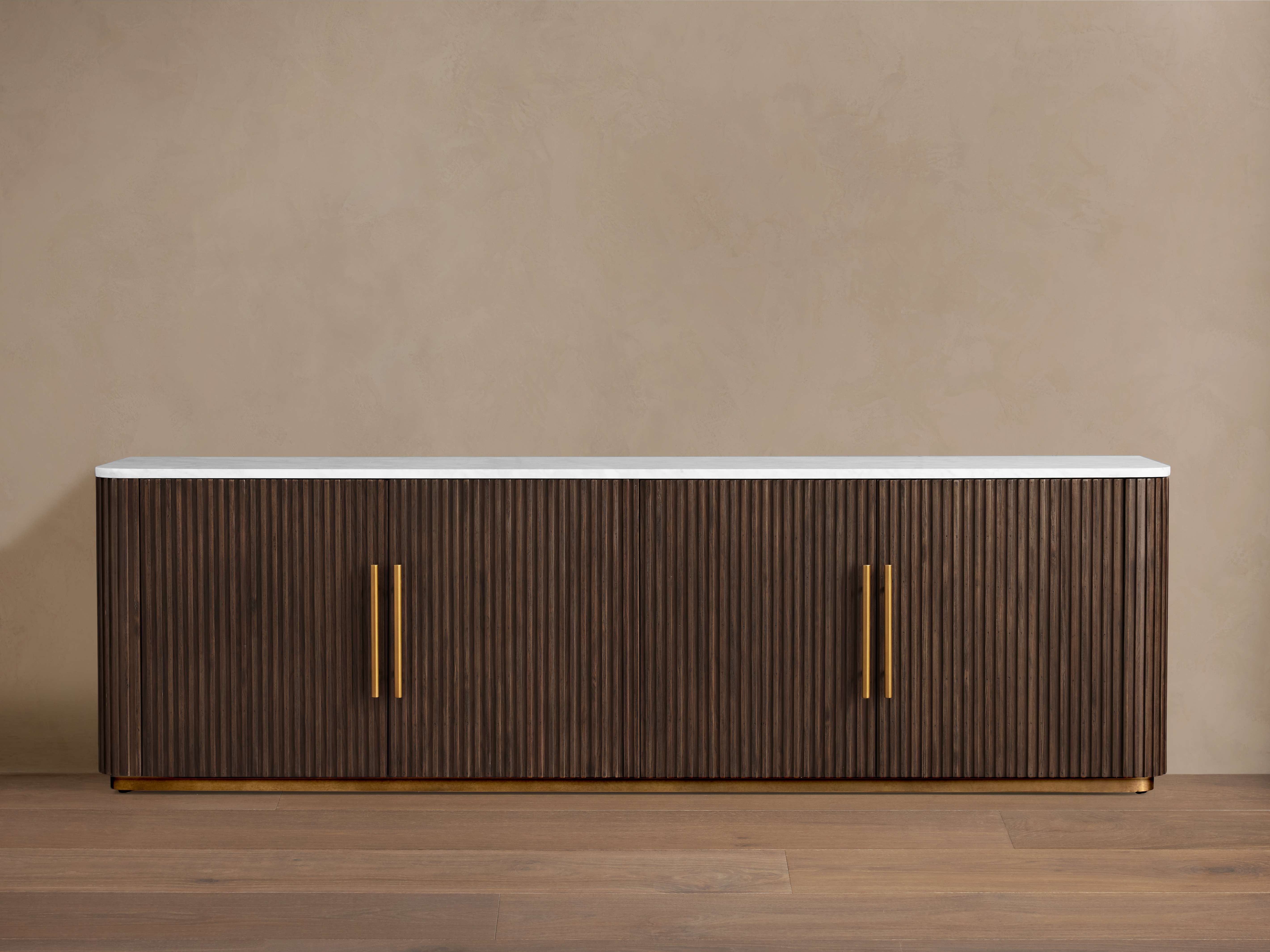 Finnley Media Console with dark ribbed wood finish, white marble top, and light brass vertical handles, placed on wooden floor against a beige wall.