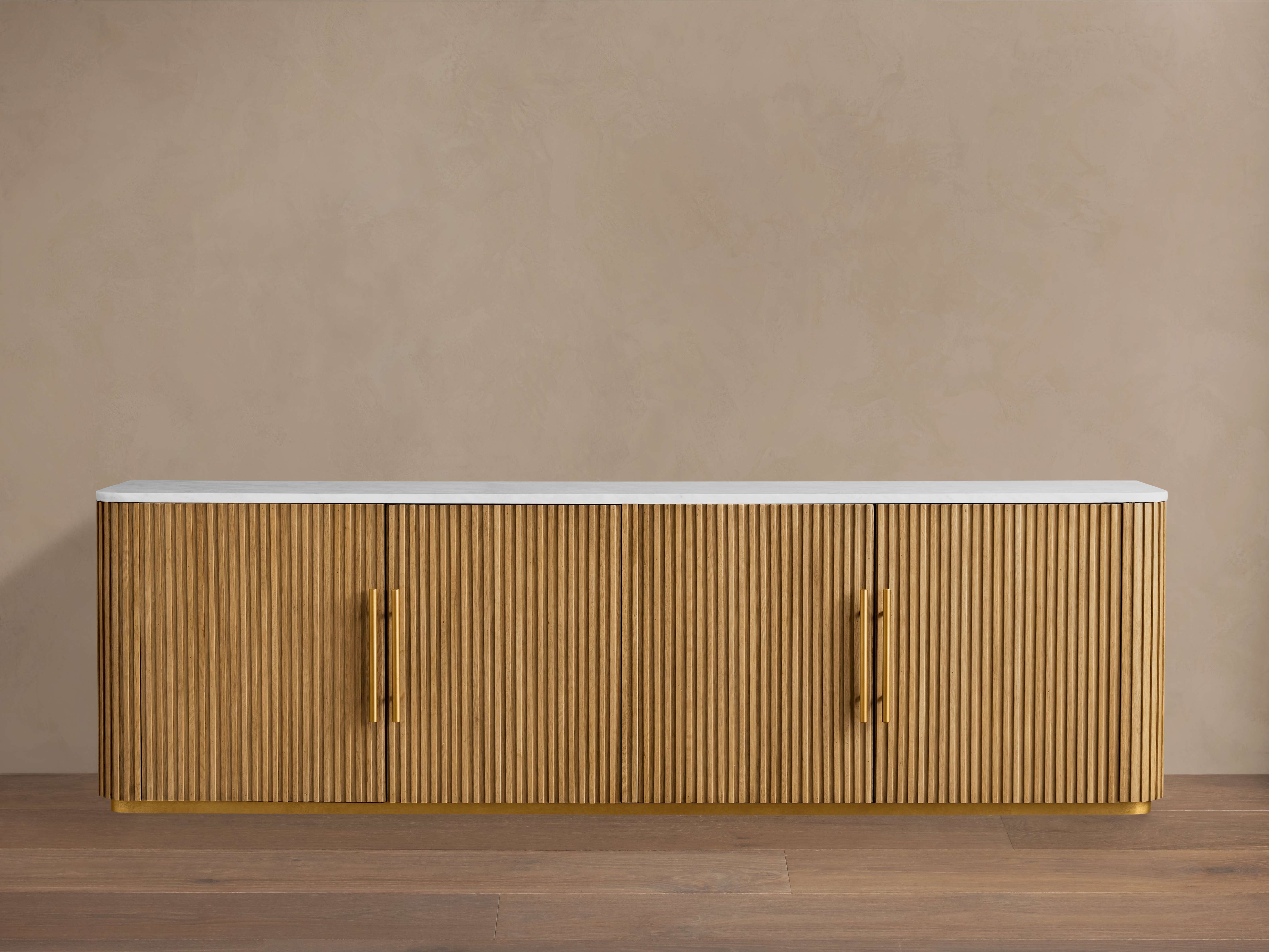 Finnley Media Console in honey finish with vertical grooves, four doors, brass-finished pulls, and a white stone top.