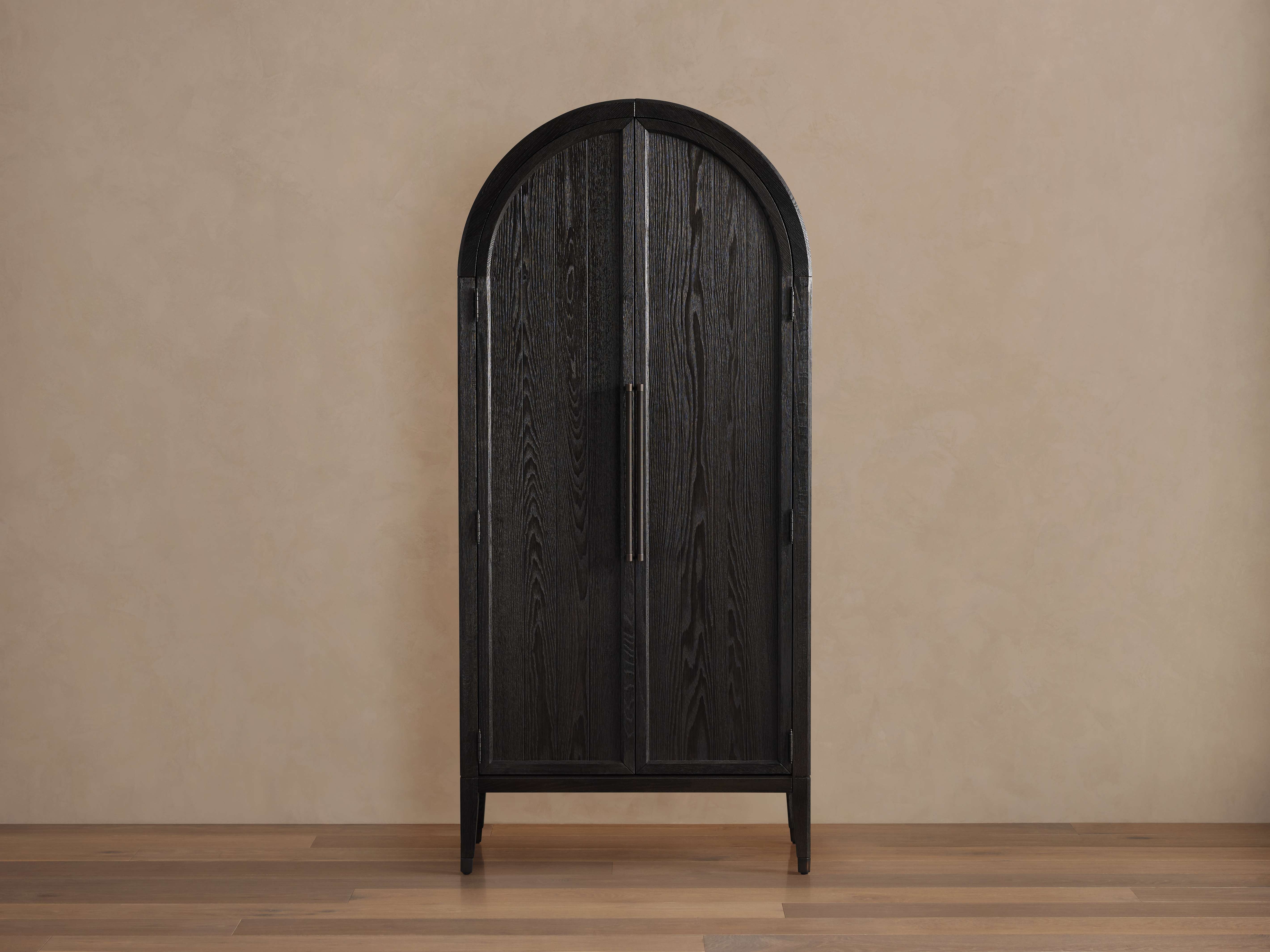 Hattie Cabinet with arched top, dark oak finish, vertical wood grain, and brass hardware, standing on a wooden floor.