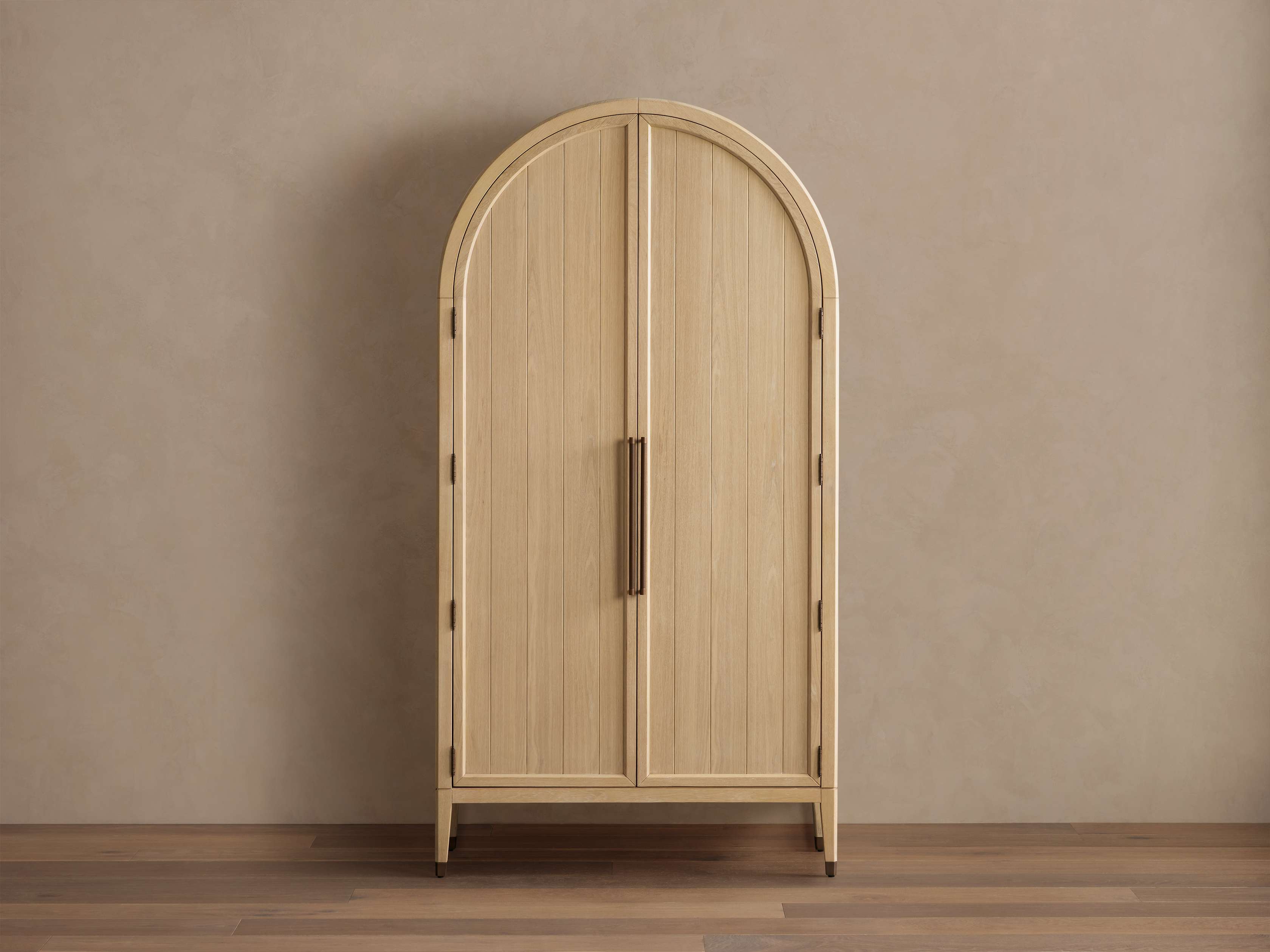 Oak wood Hattie Cabinet with an arched top, reeded hardware, and brass-capped feet against a beige wall and wooden floor.