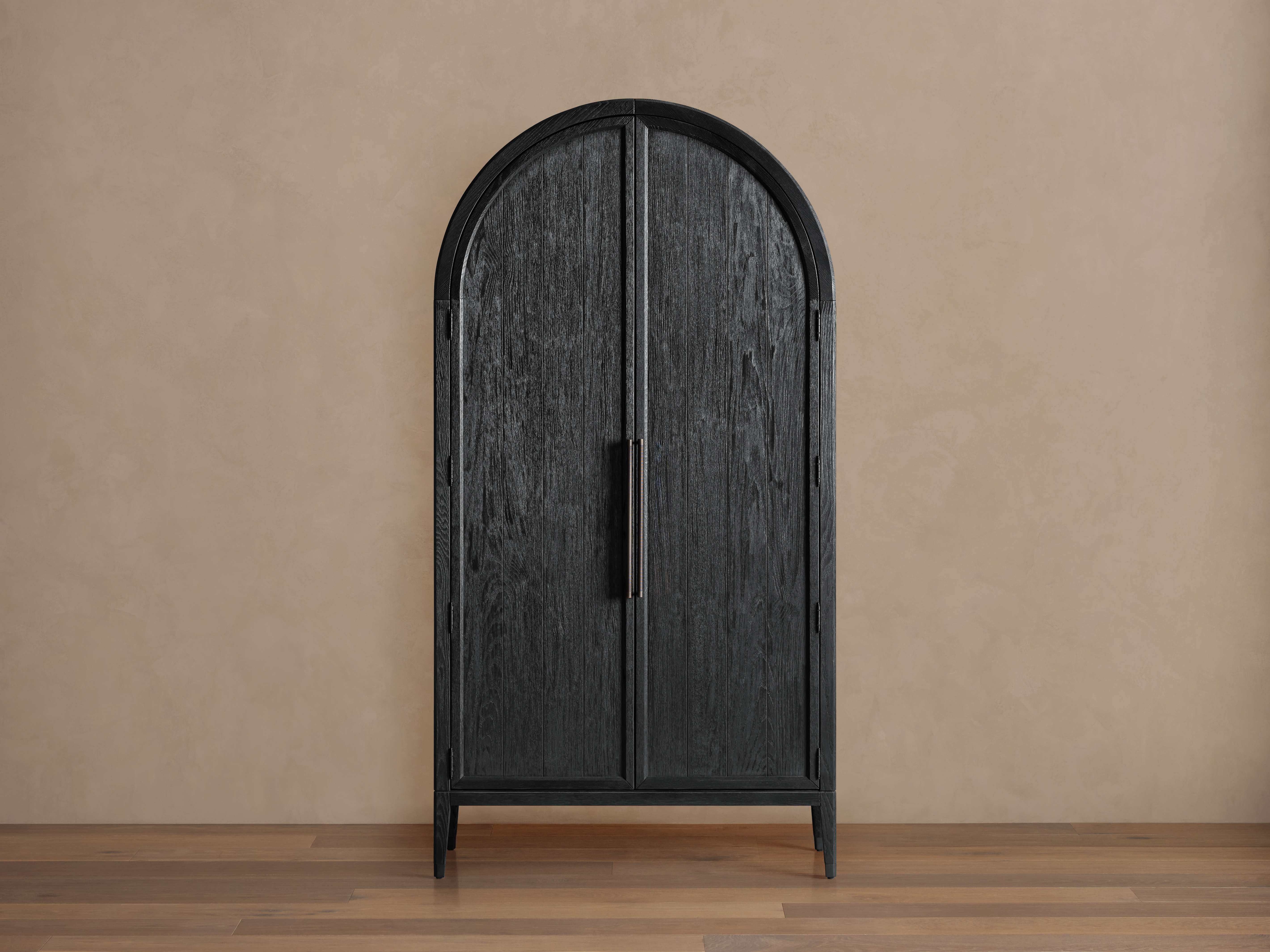 Arched black Hattie Cabinet made of oak wood, with closed doors and exposed hinges, on a wooden floor with beige wall backdrop.