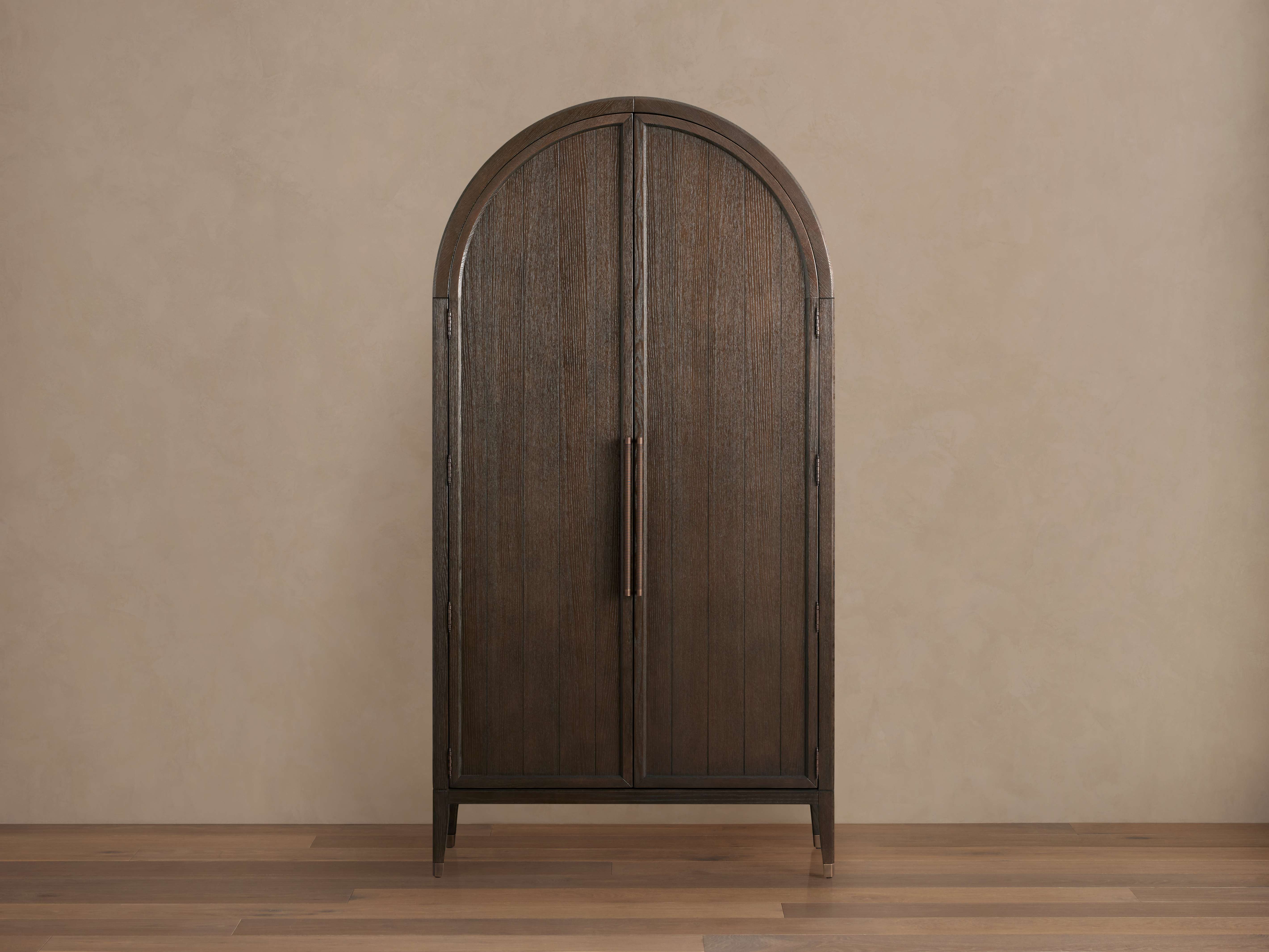 Arched Hattie Cabinet with dark oak finish, two doors with exposed hinges, reeded hardware, set on a wooden floor against a beige wall.