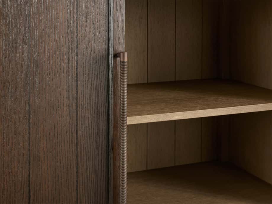 A close-up of the Hattie Cabinet made from solid oak with a dark finish. Features reeded hardware on the door and visible wood grain. Inside, shelves are visible against a shiplap back panel.