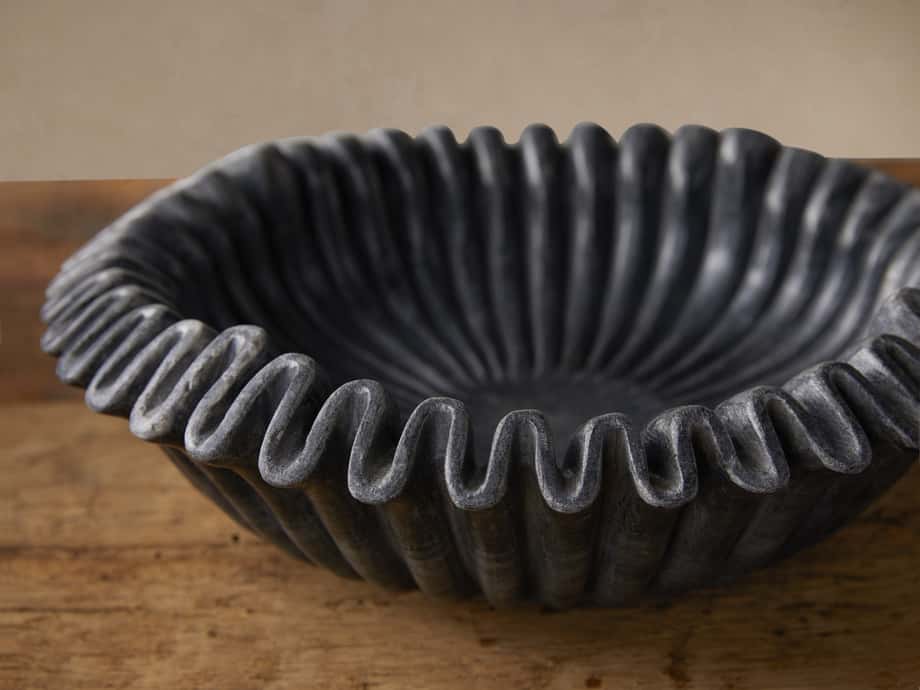 Black Marble Ruffle Bowl, close-up. Dark grey, wavy, sculpted marble rim and ribbed interior on a rustic wooden surface.