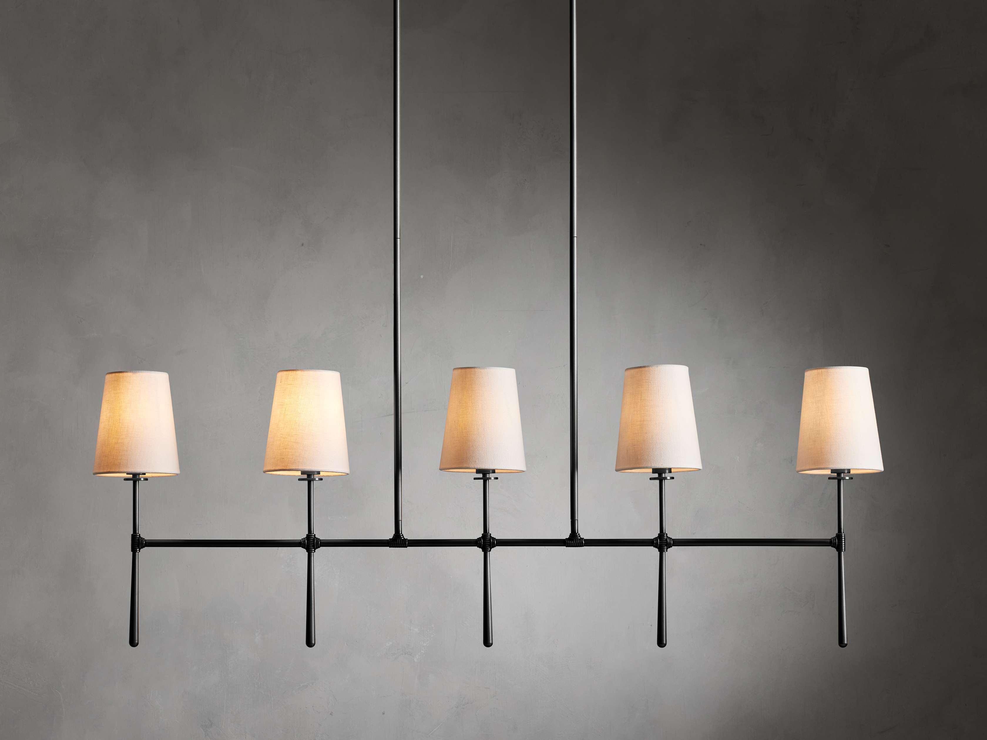 Rhodes Linear Chandelier with five black arms, each holding a tapered linen shade. Bronze plated finish against gray wall.